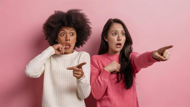 Picture of two shocked girls in sweaters pointing and looking away over white bachground