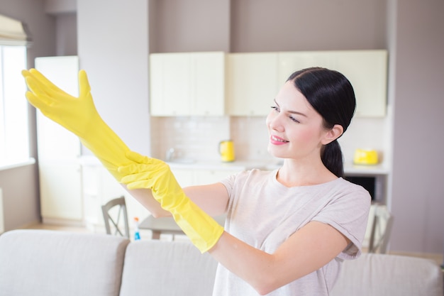 A picture of woman stands and puts on yellow glove on right hand. She looks at that and smiles. Girl stands in front of sofa in studio apartment.