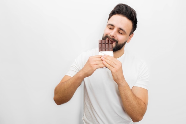 Picture of young bearded man eating delicious chocolate. He bites it with eyes closed. 