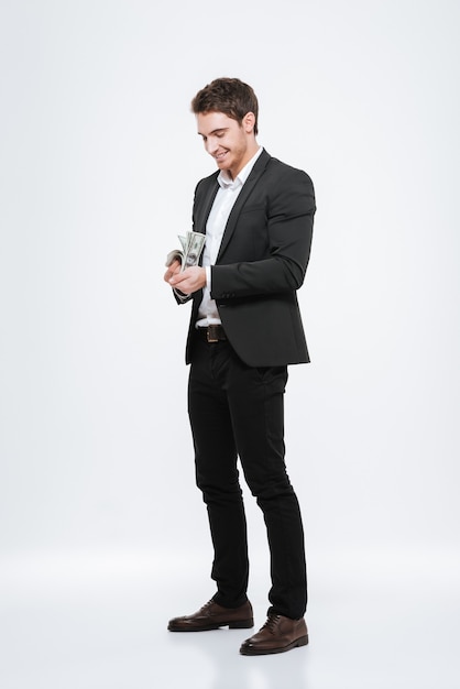 Picture of young cheerful businessman standing  holding money in hands. Isolated over white wall. Look at money.