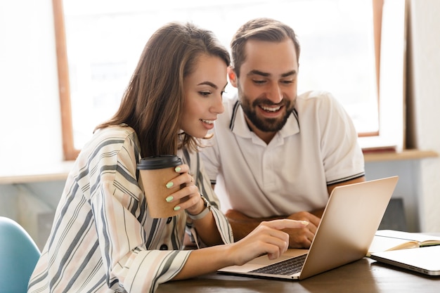 若い陽気なカップルの同僚の写真は、お互いに話しているオフィスの屋内でラップトップコンピューターを使用しています。