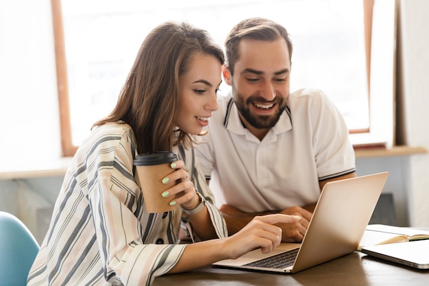 Picture of a young cheerful couple colleagues work with laptop computer indoors in office talking with each other.