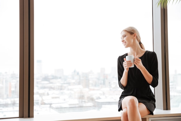 Picture of young cheerful lady worker sitting in office near window while drinking hot tea. Looking at window.