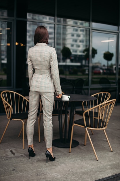 Picture of young lovely caucasian female with dark hair in stylish suit, white shoes backwards