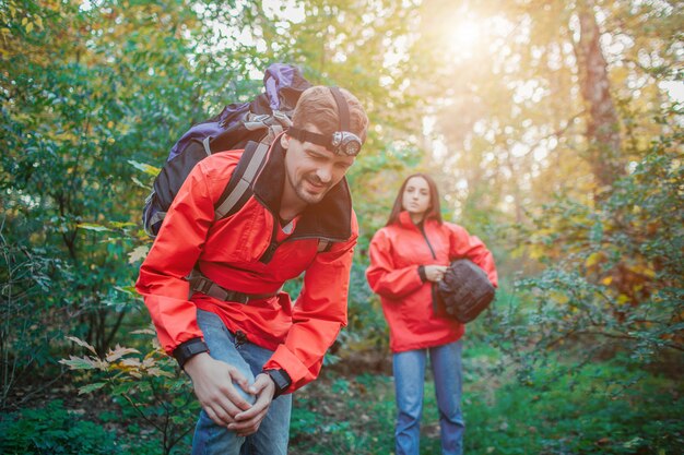 Photo picture of young man holding hands on knee. he has pain there. guu suffer. young woman stands behind him and look. she holds sleeping bag. guy has backpack on back.