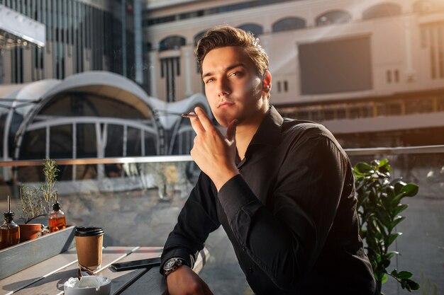 Picture of young man sitting at table and smoking. He look up to right. Guy lean on table. He sit outside. Sun is shining.