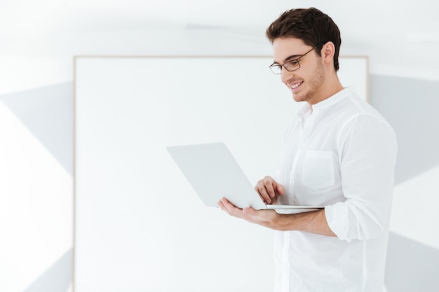 안경을 쓰고 흰 셔츠를 입고 큰 보드 근처에서 노트북 컴퓨터를 사용하는 젊은 남자의 사진. 노트북 보세요.