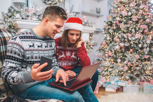 Picture of young man and woman sitting and looking at laptop. Guy has it on his nap. Also man holds phone in right hand. Couple looks concentrated.