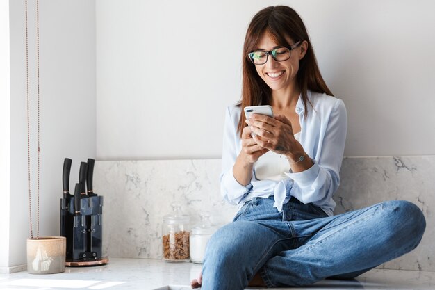 Picture of a young positive smiling happy pretty business woman sitting indoors at home at the kitchen using mobile phone chatting.