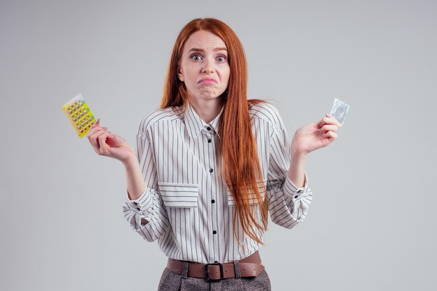 Picture of young redhead be lost in thought businesswoman in striped shirt with one pack of condom and birth control pills choosing thinking white background studio