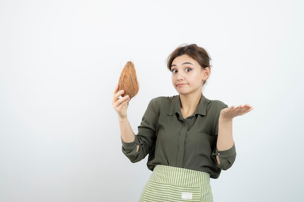 Picture of young woman in apron holding a coconut against white wall . High quality photo