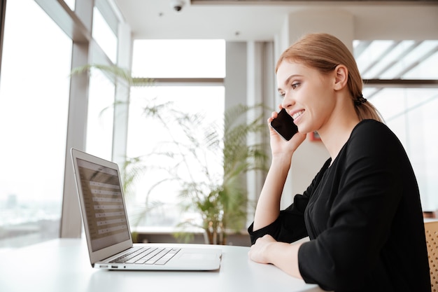 Picture of young woman worker sitting in office while using laptop computer and talking by her phone. Looking at computer.