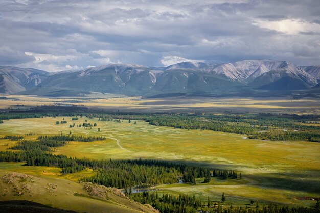 Picturesque green valley in sunlight against snowy mountain range under cloudy sky on summer or autumn day.