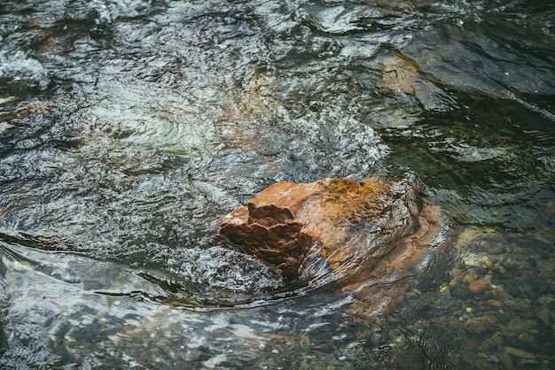 Живописный фон природы с ярким оранжевым камнем в прозрачной воде горной реки. Красочное каменистое дно под чистой водой. Красивый фон природы с оранжевым валуном в горном ручье.