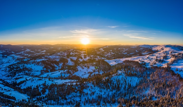 Picturesque winter panorama of mountain hills covered with snow and fir trees on a sunny clear day with the sun and blue sky. Pristine Nature Beauty Concept. Copyspace