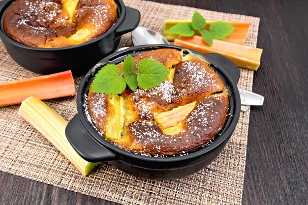 Pie klafouti with rhubarb in black brazier, mint and rhubarb stems on wicker napkin on wooden plank background