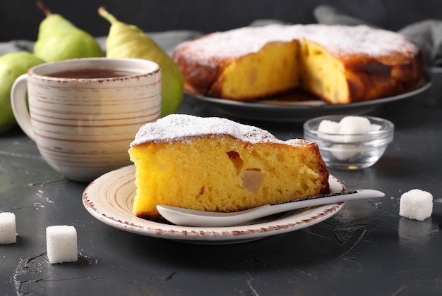 Photo pie with pears with a cut piece and a cup of tea on a dark background.