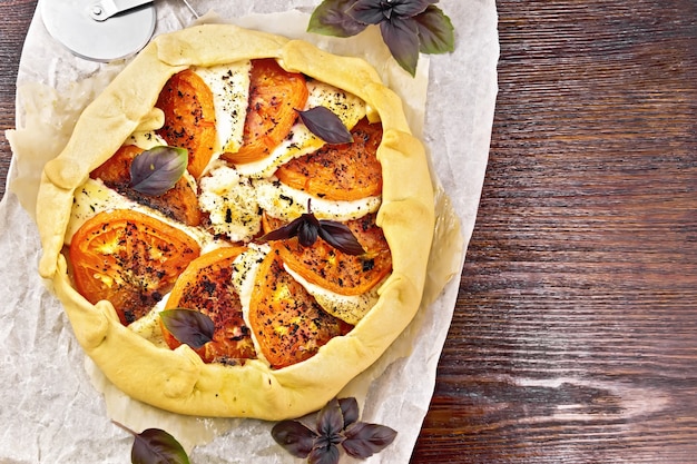 Pie with tomatoes, curd cheese and purple basil on parchment on a wooden board background from above