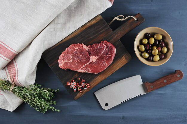 Piece of beef on a wooden Board, a knife for meat, olives and thyme 