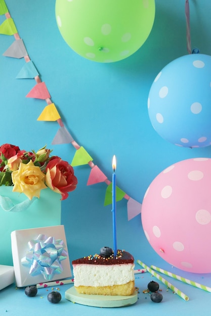 Кусок ягодного торта с горящей свечой, букет свежих роз, подарки, воздушные шары