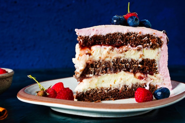 A piece of cake chocolate biscuit with cream cheese filling on dark blue background close up Berries on pink cream decoration High quality photo