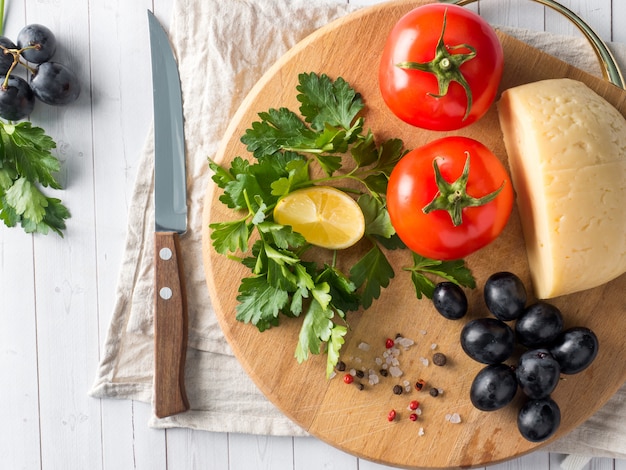 Pezzo di formaggio con prezzemolo, uva pomodori su un tagliere con un coltello.