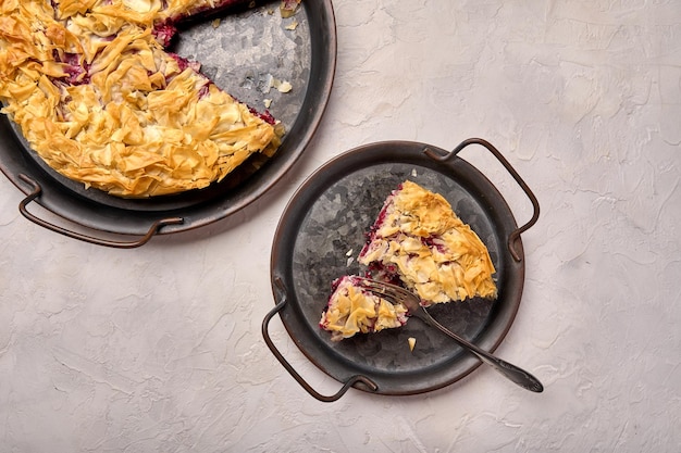 Piece of cherry pie from filo dough and fork on rustic metal tray on light background top view