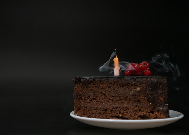 Piece of chocolate birthday cake with one extinguished candle\
on white plate on deep black backgroun