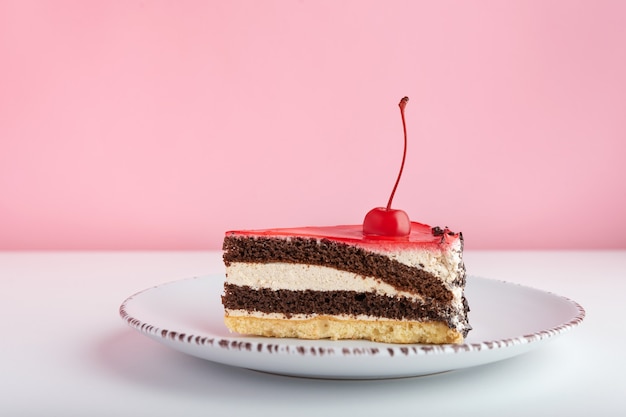 Piece of chocolate vanilla cake with maraschino cherry. Side view cake on white plate on pink background.