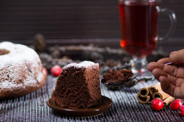Кусок рождественского шоколадного бисквита с сахарной пудрой на деревянном столе со стаканом красного чая и палочками корицы