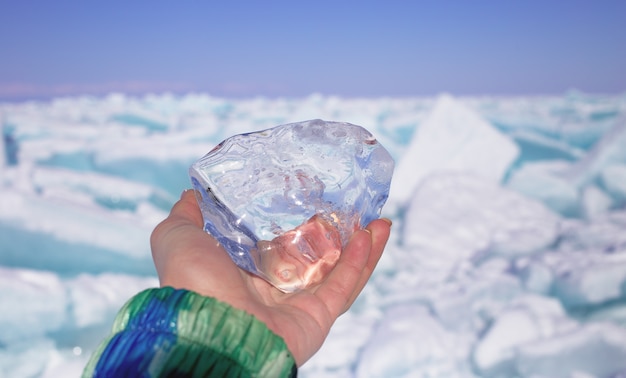 A piece of crystal transparent ice in the hand against frozen lake in sunny day