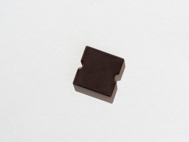 A piece of dark chocolate bar on a white paper background. A hard shadow. Sweet and high-calorie product. Top view, flat lay