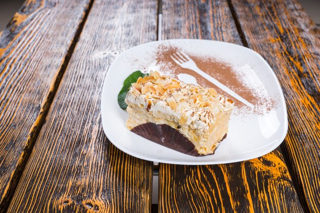Pezzo di torta alla crema decadente condita con noci servita su un piatto bianco e guarnita con foglie di menta e spolverata di cacao sulla superficie del tavolo in legno
