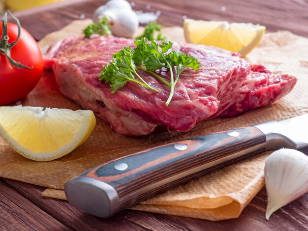 A piece of fresh raw beef lies on parchment surrounded by spices, herbs and vegetables. The knife is lying nearby. Side view. Food composition