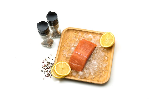 A piece of fresh salmon with lemon on ice in a wooden square plate. Salt and spices.