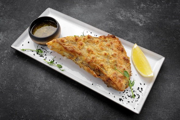 A piece of fried flounder fish with lemon on a plate on a dark background