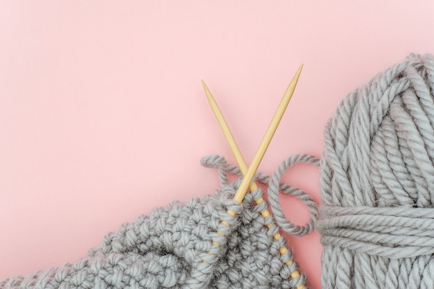 Photo piece of grey knitted fabric on bamboo wood needles with ball of yarn on pink background