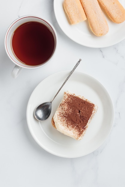 Piece of Homemade Tiramisu cake dessert and coffee with savoiardi as ingredients on a white marble background