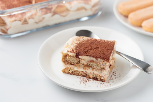 Piece of Homemade Tiramisu cake dessert with savoiardi as ingredients on a white marble background