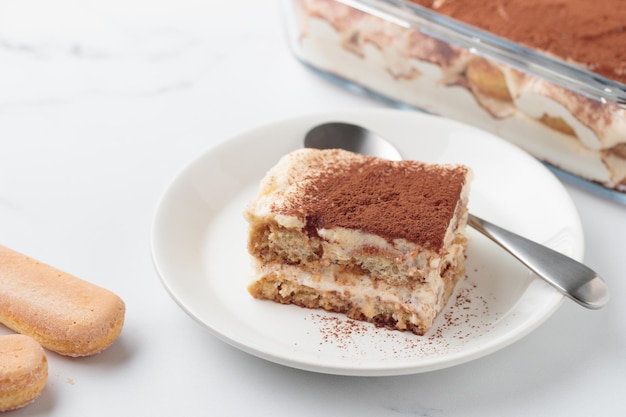 piece of homemade tiramisu cake dessert with savoiardi as ingredients on a white marble background