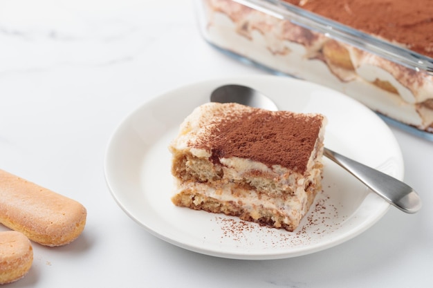 Piece of Homemade Tiramisu cake dessert with savoiardi as ingredients on a white marble background