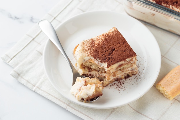 piece of homemade tiramisu cake dessert with savoiardi as ingredients on a white marble background