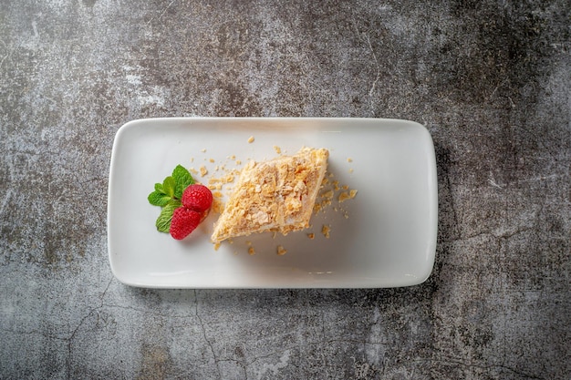 Piece of layered cream cake with raspberries and mint in a white plate against a gray stone table in a restaurant, a delicious breakfast