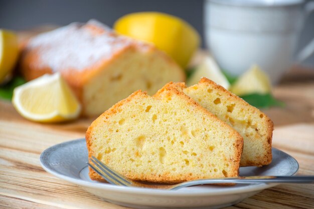 Piece of lemon cake on plate on rustic wooden board with full pie, lemons and cup on background.