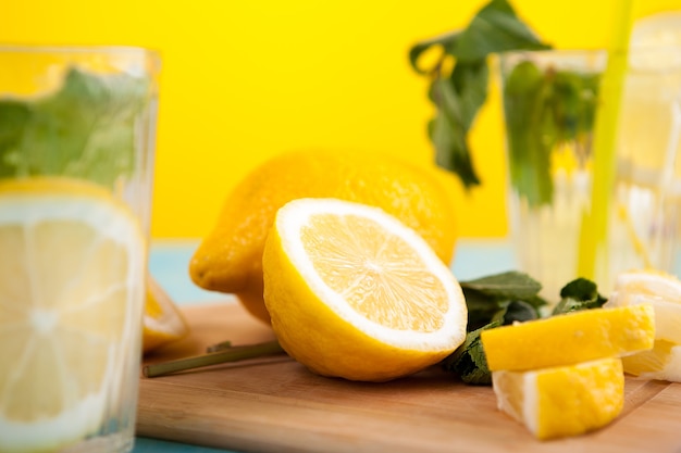 A piece of lemon cutted in two on wooden board next to a full lemon and two glasses with natural homemade lemonade