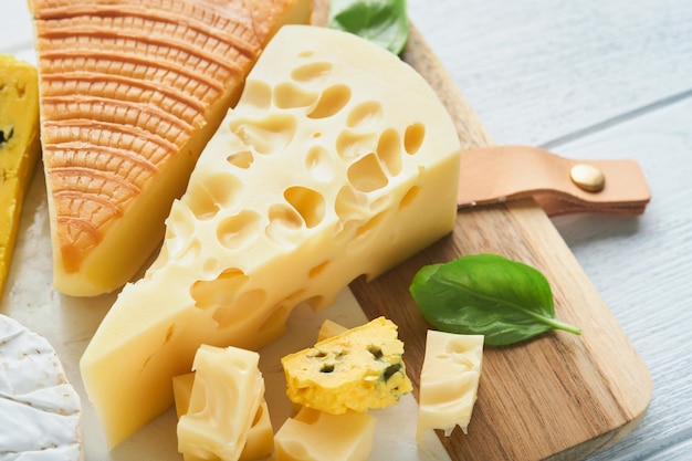 Piece of maasdam cheese and camembert smoked cheese blue cheese\
parmesan brie cheese with basil and pepper on white marble board\
white wooden old background set or assortment cheeses top view