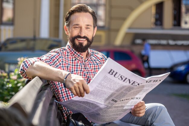 Piece of news. Delighted smiling man reading a daily newspaper while resting outdoors