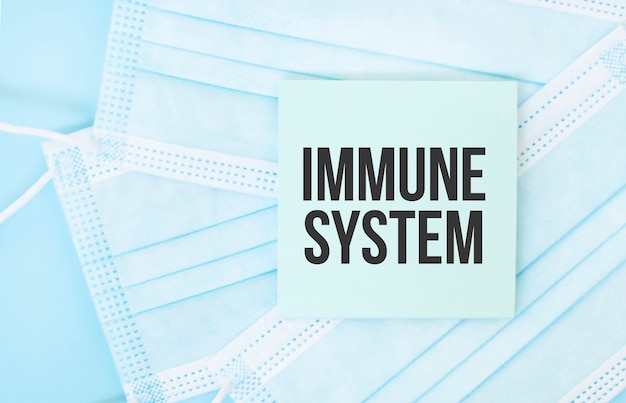 青い医療用マスクの山にIMMUNESYSTEMというフレーズが書かれた一枚の紙。コロナウイルスパンデミックの概念