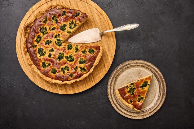 Piece of pie on plate and quiche tart with broccoli and tuna on round wooden cutting board with