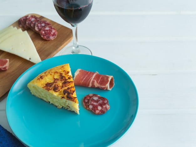 Piece of potato omelette with ham and sausage on a blue plate with a glass of wine on a white table.